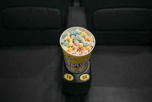 A tub of colorful popcorn resting on a seat armrest in a movie theater.