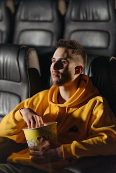 A young man in a yellow hoodie watches a movie intently while holding popcorn.