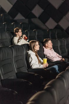 Three young adults seated in a movie theater enjoying a film. Holding snacks and drinks.