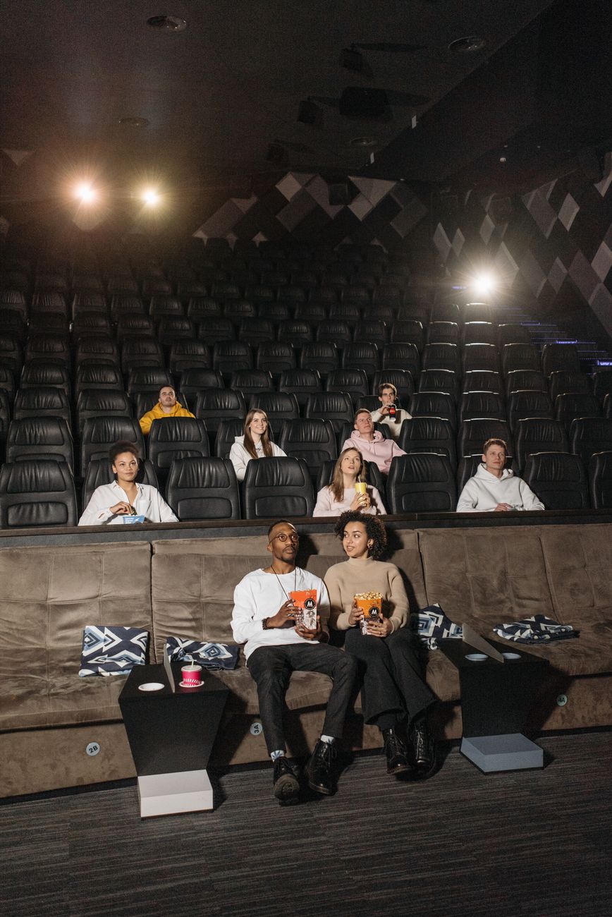 Un groupe de personnes est assis dans une salle de cinéma sombre. Au premier plan, un homme et une femme mangent du pop-corn sur un grand canapé. D'autres spectateurs sont installés sur les sièges derrière.