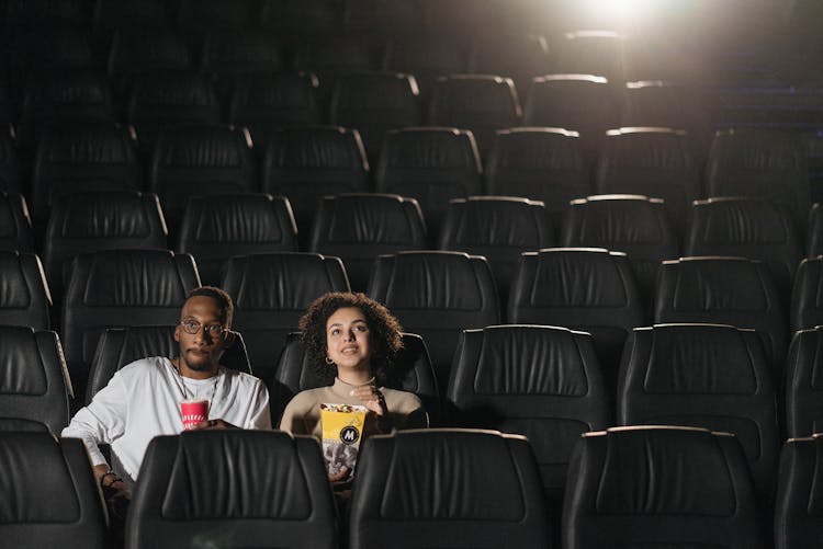 A Couple Sitting On Black Leather Seats With A Cup Of Drink And A Tumbler Of Popcorn