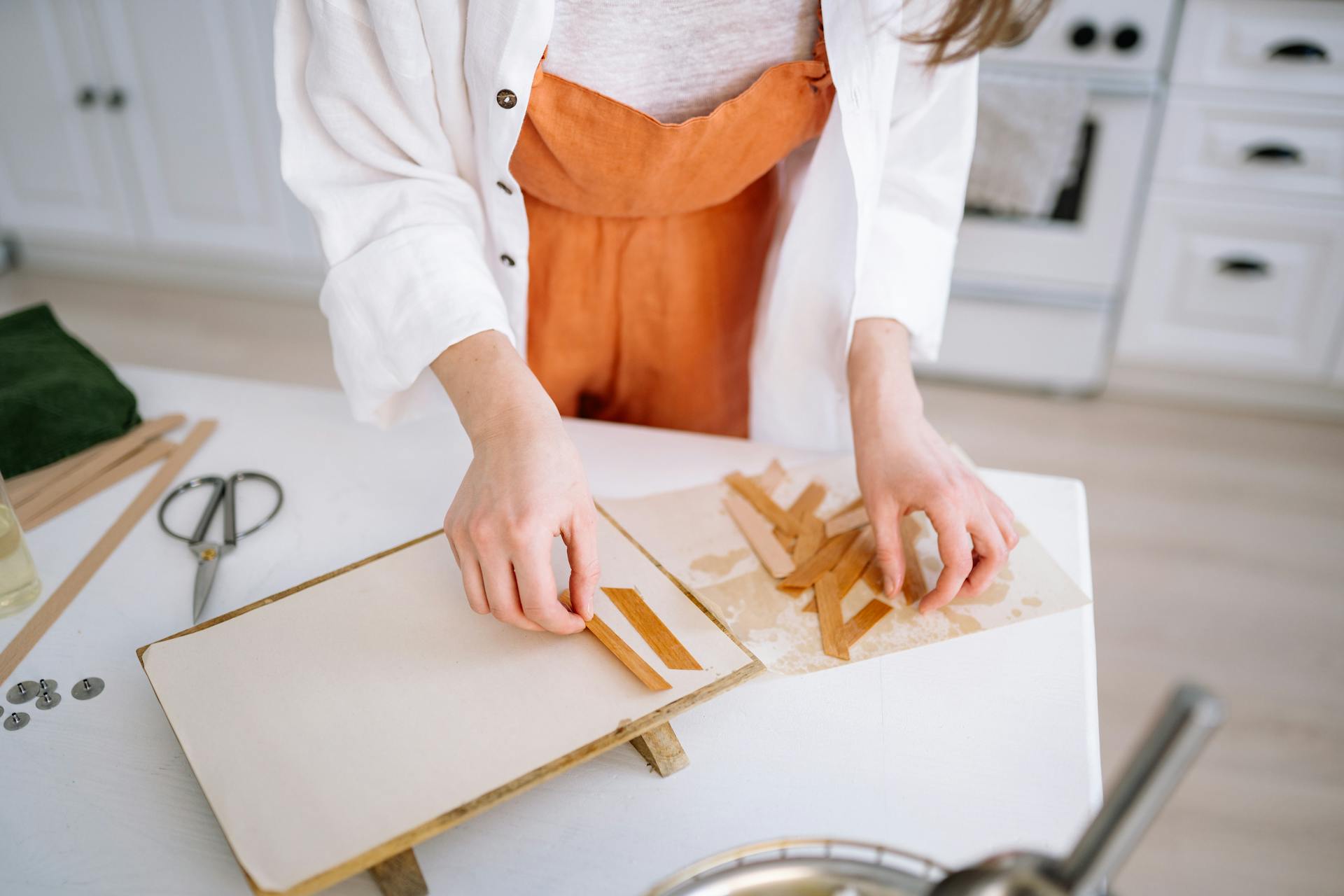 A woman in a kitchen arranging wooden strips for crafting on a white table.