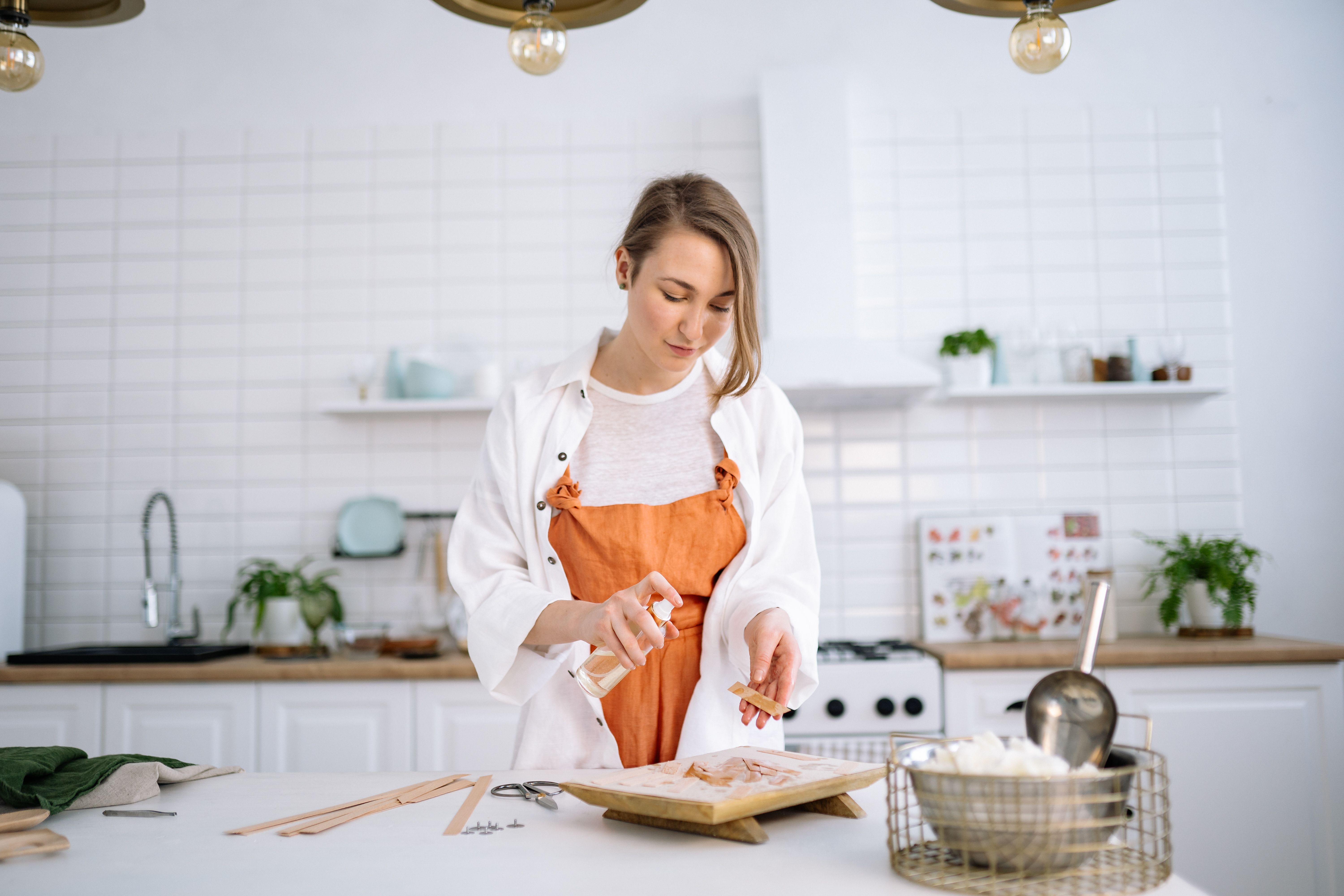 A woman crafting candles in a modern kitchen workshop, wearing an apron, spray bottle in hand.