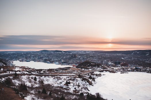 Grassy shore covered with snow near frozen water against hill with residential houses in coastal area on cold winter day