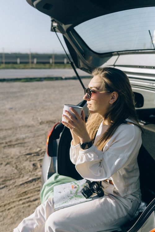 Free Woman sits in car trunk drinking coffee while holding a map and wearing sunglasses. Stock Photo