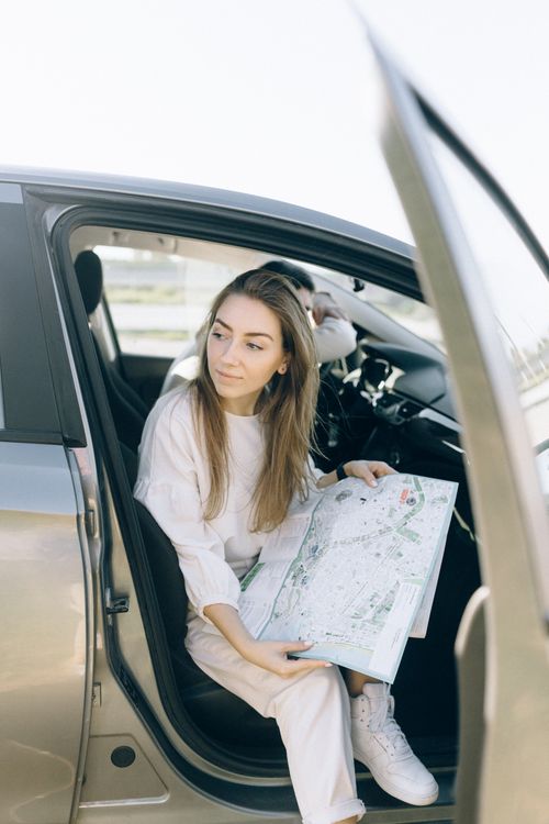 Free Young woman sitting in car holding a map, planning her journey with the door open. Stock Photo