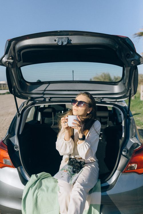Free Woman sitting in car trunk enjoying coffee outdoors on a sunny day. Stock Photo