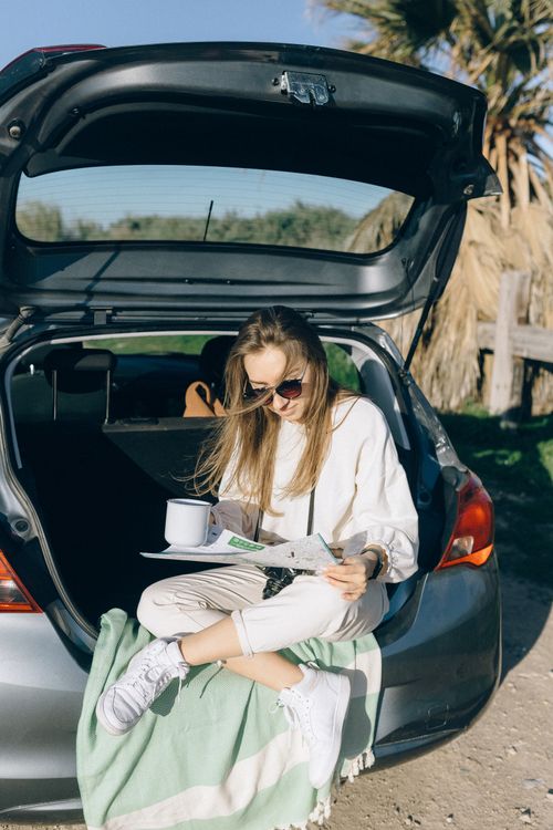 Free Young woman with sunglasses reading a map and enjoying a break in a car trunk outdoors. Stock Photo