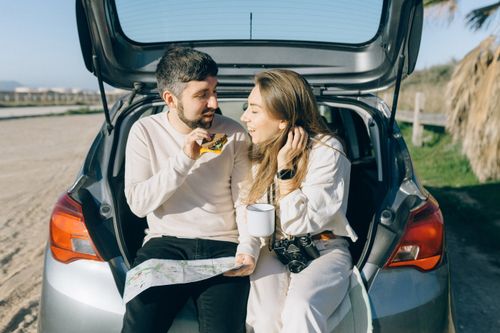 Free Couple enjoying a road trip, sitting in a car trunk, sharing a snack on a sunny day outdoors. Stock Photo
