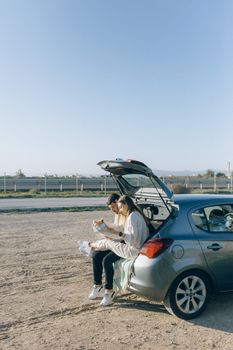 A couple sitting on a car trunk, enjoying a road trip outdoors on a sunny day.
