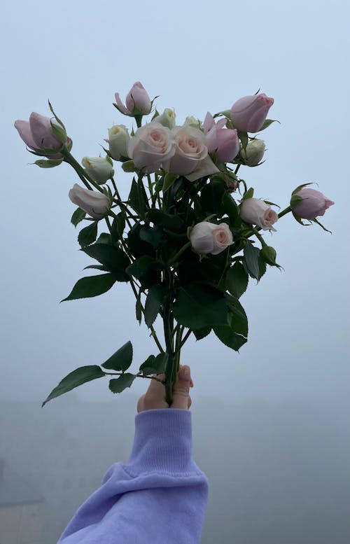 Unrecognizable female with bouquet of blooming roses with green leaves in hand standing on street in rural area in misty weather