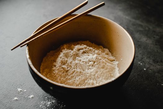 Close-up of flour in a bowl with chopsticks on a dark surface, perfect for culinary themes.