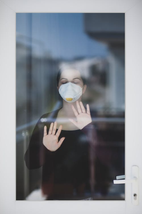 Photo of a Woman with a White Face Mask Touching a Glass Window