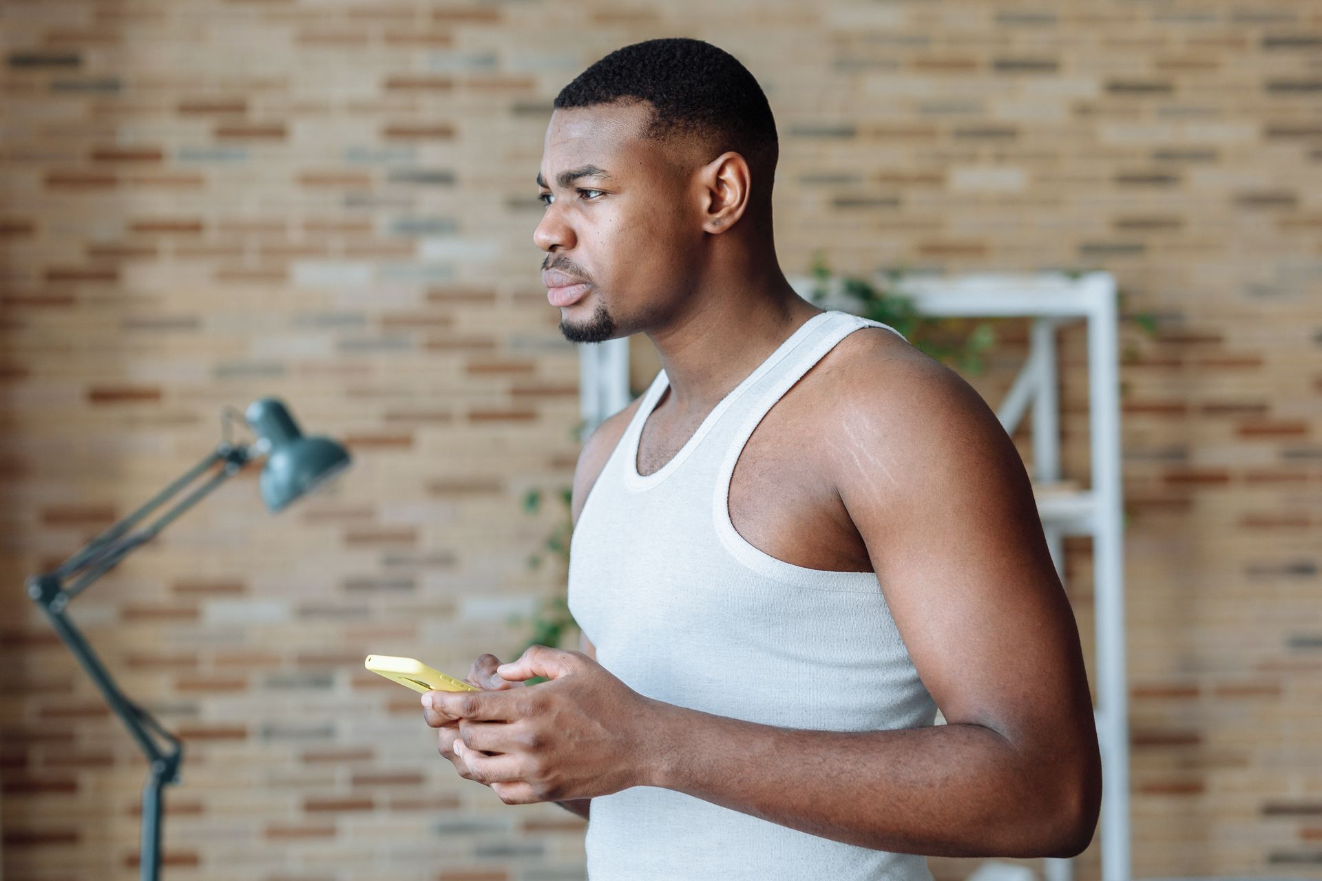 A young African American man in a white tank top uses a smartphone indoors.