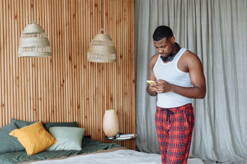 Free A man in pajama relaxes in a stylish bedroom, engaging with a smartphone. Stock Photo