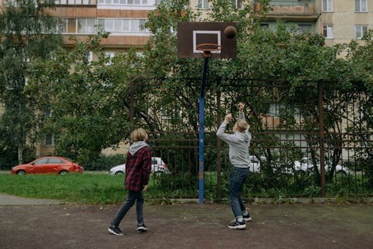 Two kids enjoying a game of basketball on an outdoor urban court, surrounded by greenery.