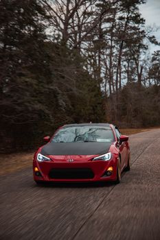 A vibrant red sports car driving on a forest road during a calm day; perfect for automotive and travel themes.