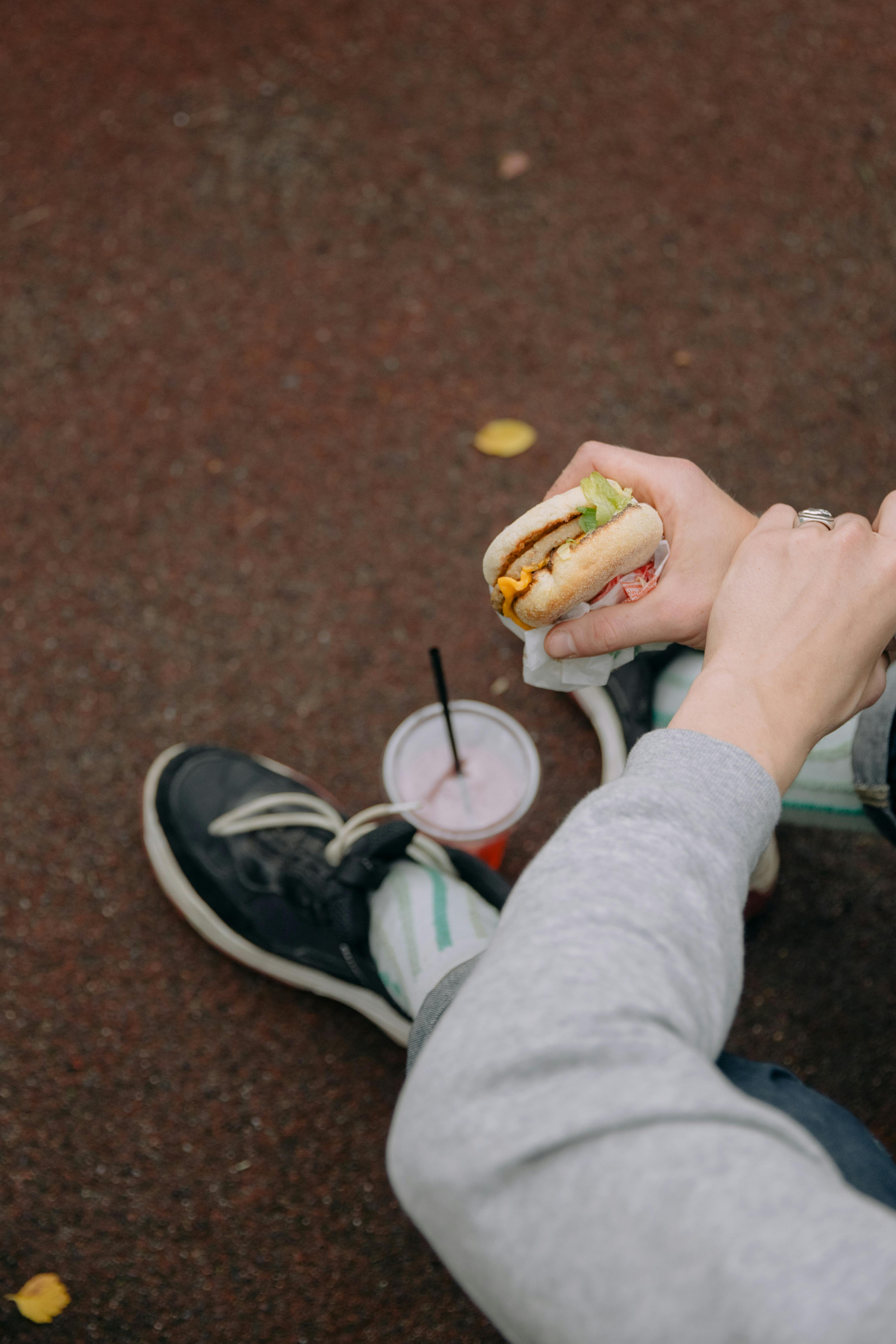 Person sitting outdoors enjoying a cheeseburger with a drink, captured from above.
