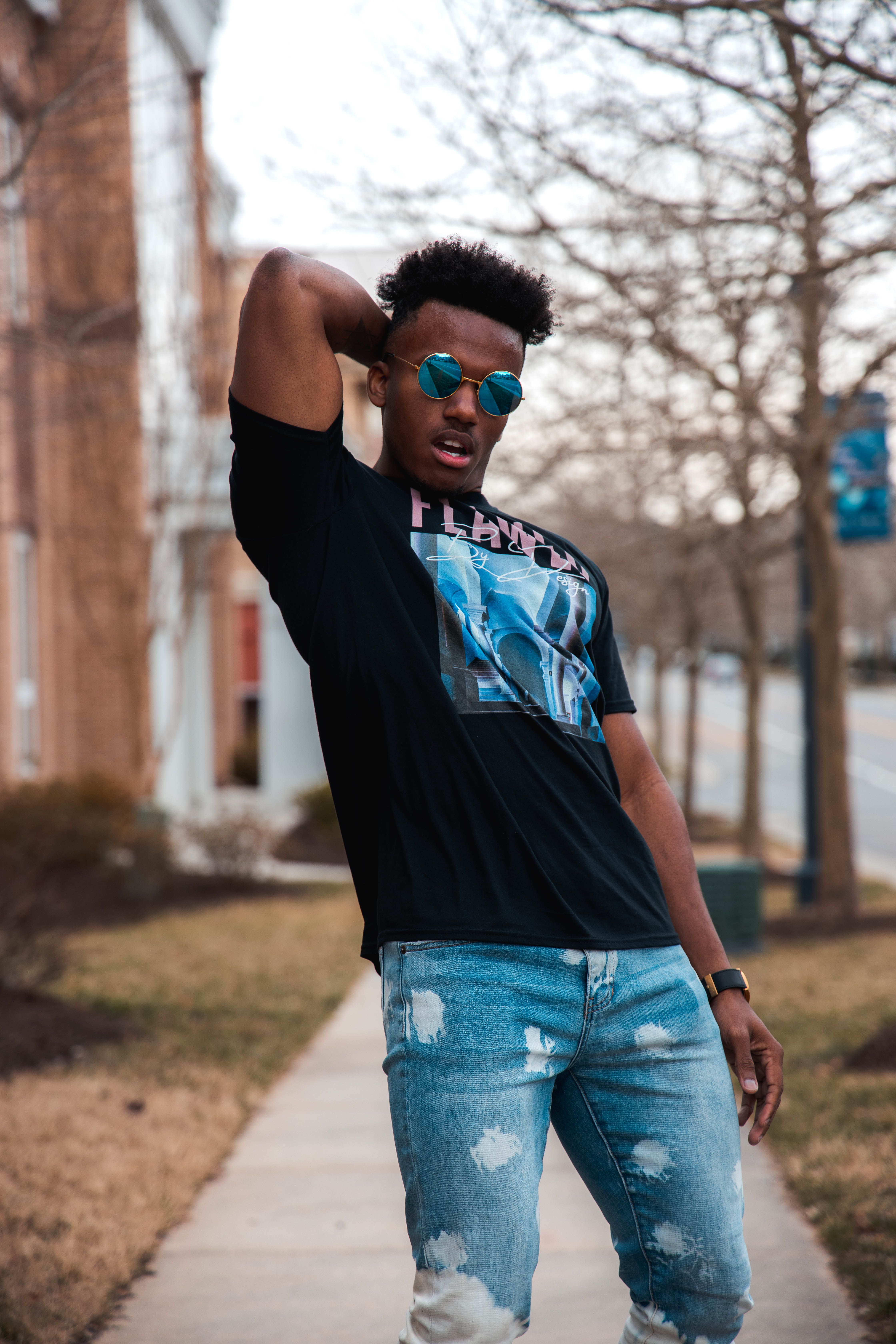 Confident young man in sunglasses and ripped jeans posing on a sunny sidewalk.