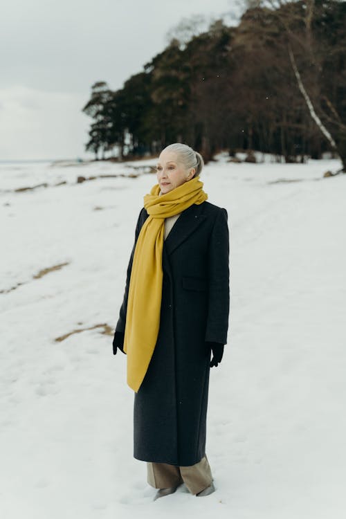 Woman in Black Coat and Yellow Scarf Standing on Snow Covered Ground