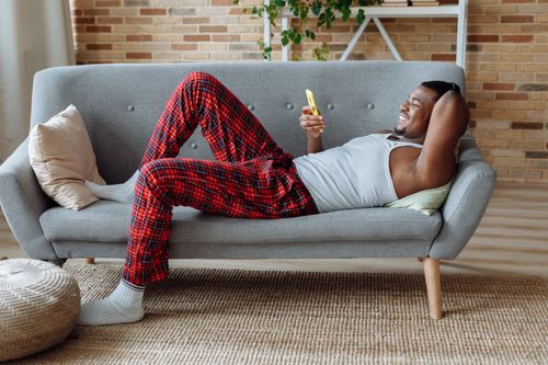 Free Cheerful young man in checked pajamas relaxing on a sofa using a smartphone indoors. Stock Photo
