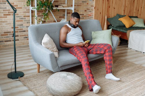 A Man in White Sleeveless Shirt Using a Cellphone while Sitting on a Sofa