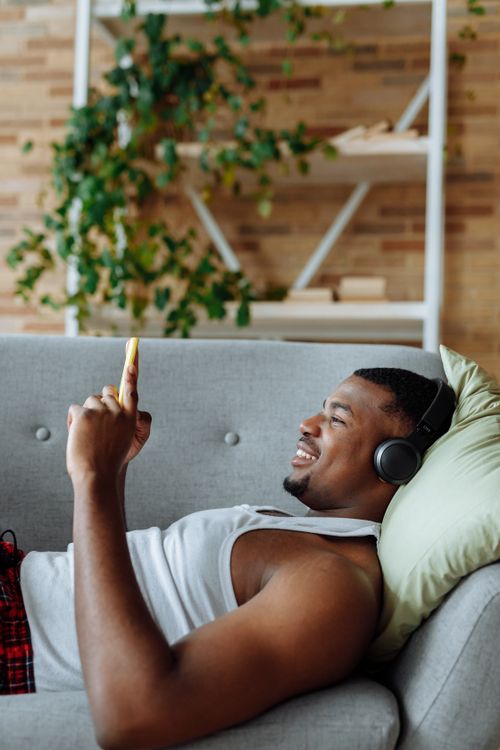Free African man lying on a sofa indoors, enjoying music with headphones and mobile phone. Stock Photo