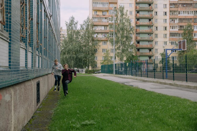 Young Men Running On The Green Grass Near The Buildings