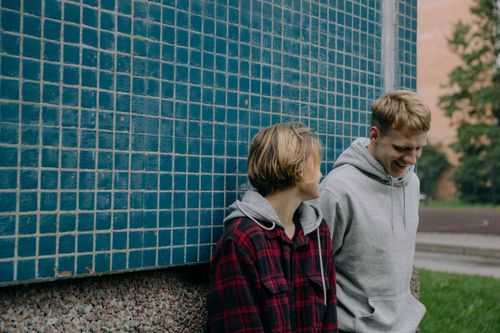 Free Two young friends in casual attire laughing against a blue tiled wall outdoors, enjoying a casual moment together. Stock Photo