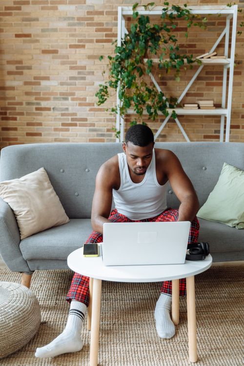 Free Man in plaid pants using laptop in a modern living room on a comfy sofa, focusing intently. Stock Photo