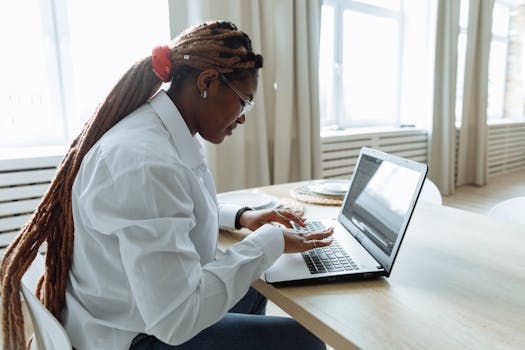 A woman with dreadlocks focuses intently on her laptop in a bright, cozy home setting.