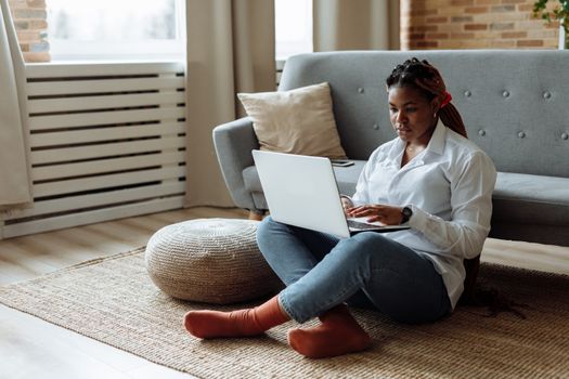 African American woman sitting on floor working from home on laptop in a cozy living room.