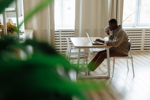 Free African American man working remotely from home using a laptop, enjoying coffee in a cozy setting. Stock Photo