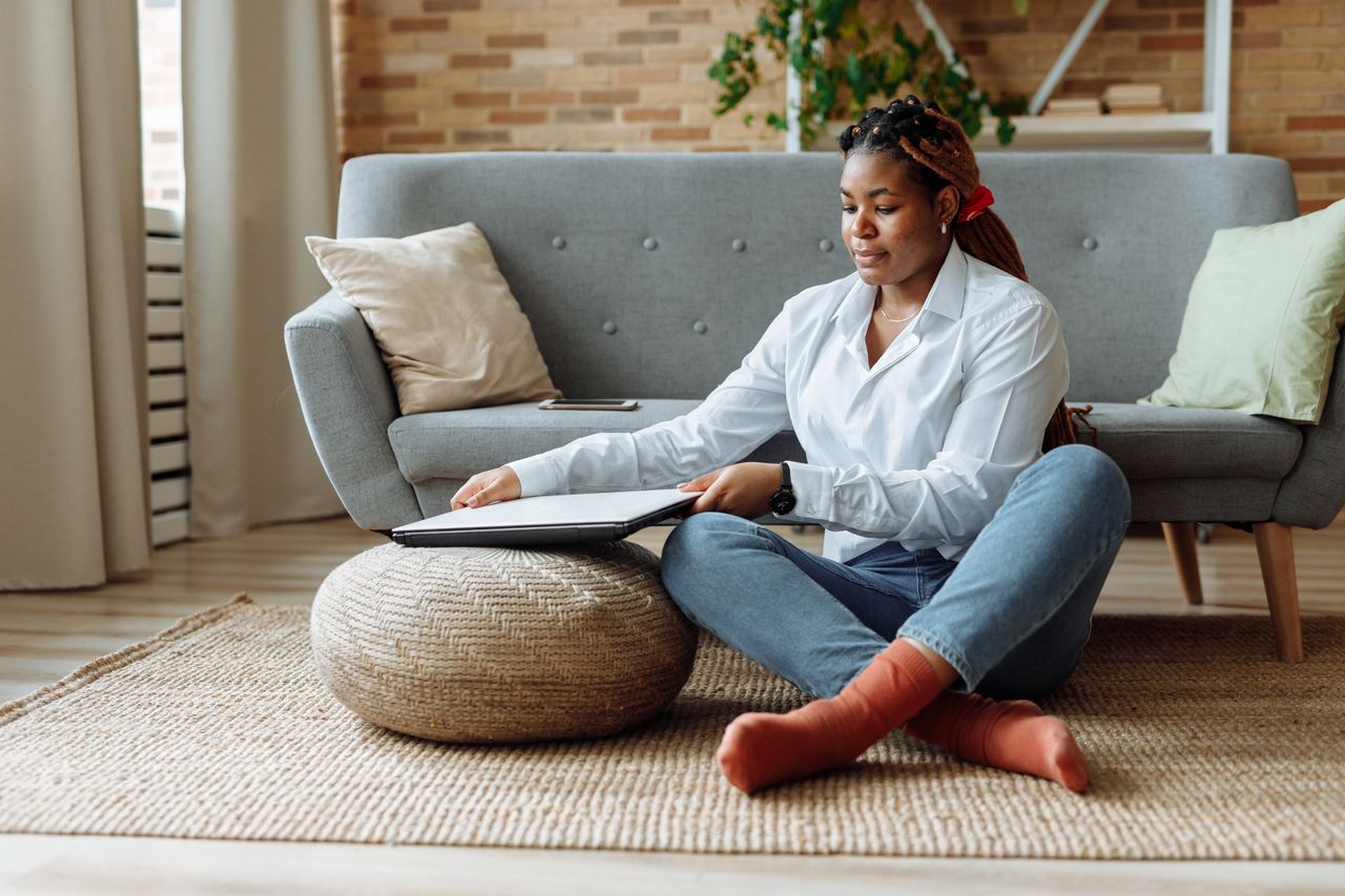 Woman using a round woven pouf as a laptop table in a living room.