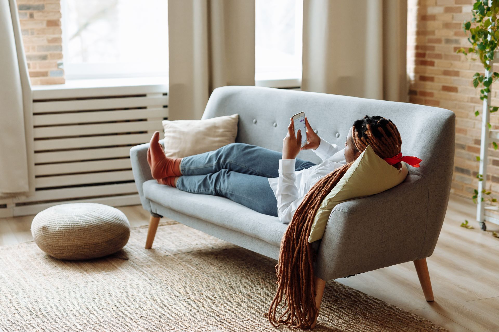 African American woman with dreadlocks relaxing and using phone on a modern sofa.