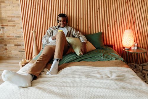 Free Young man lying on bed listening to music with headphones, smiling and relaxing indoors. Stock Photo