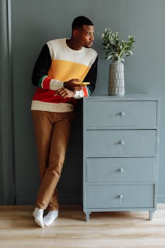 A fashionable man wearing a colorful sweater uses his cellphone while leaning against a cabinet indoors.