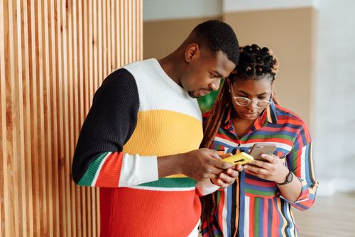 Free A young couple stands together indoors, focused on their smartphones, demonstrating connectivity and modern communication. Stock Photo