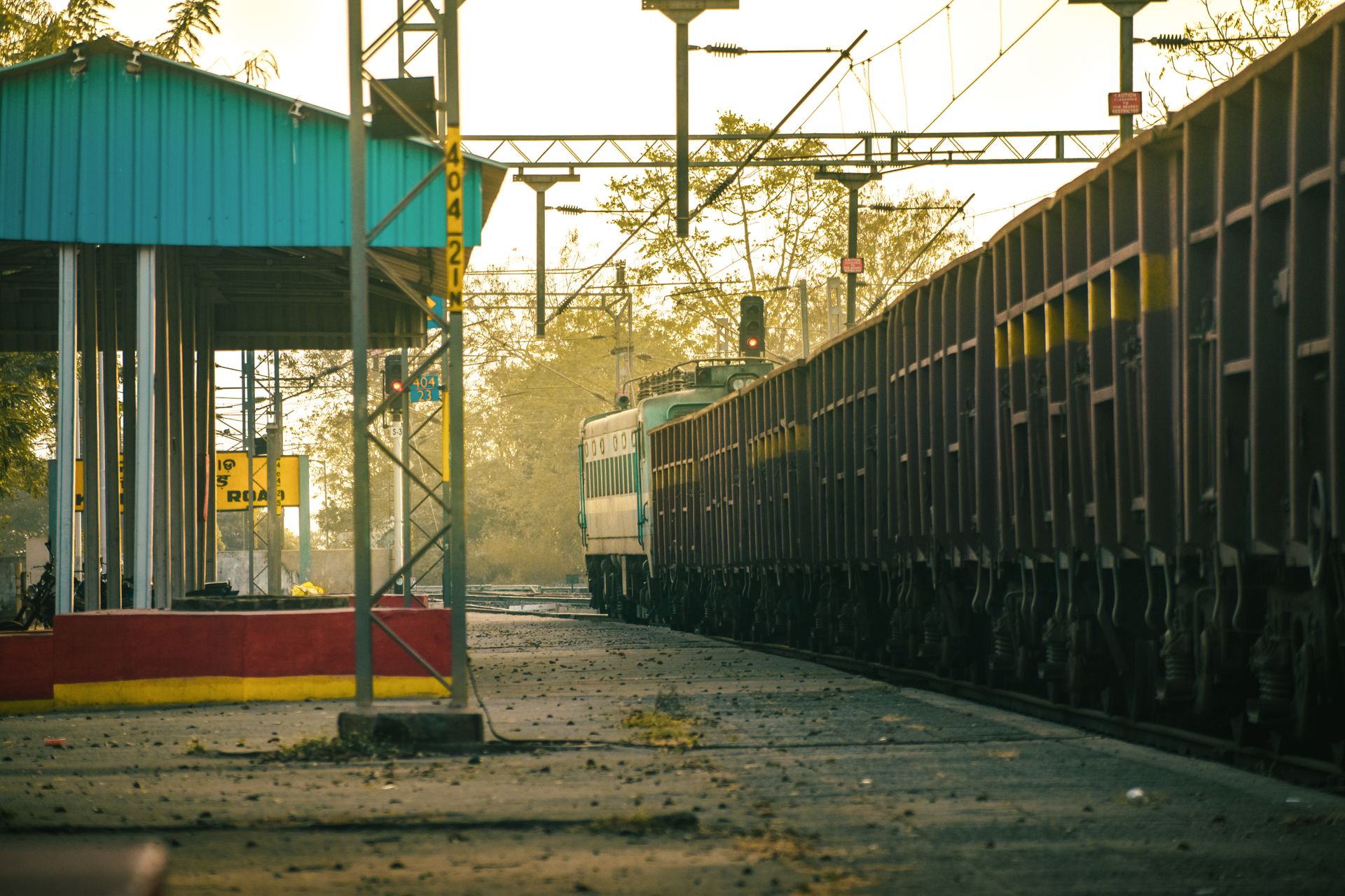 A freight train arrives at an Indian railway station platform during a calm day.