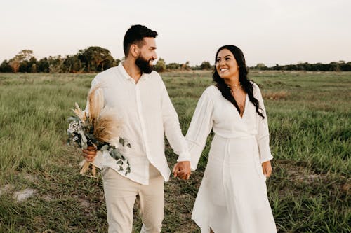 Free Smiling loving couple with bouquet of meadow grass strolling in grassy lawn and looking at each other Stock Photo