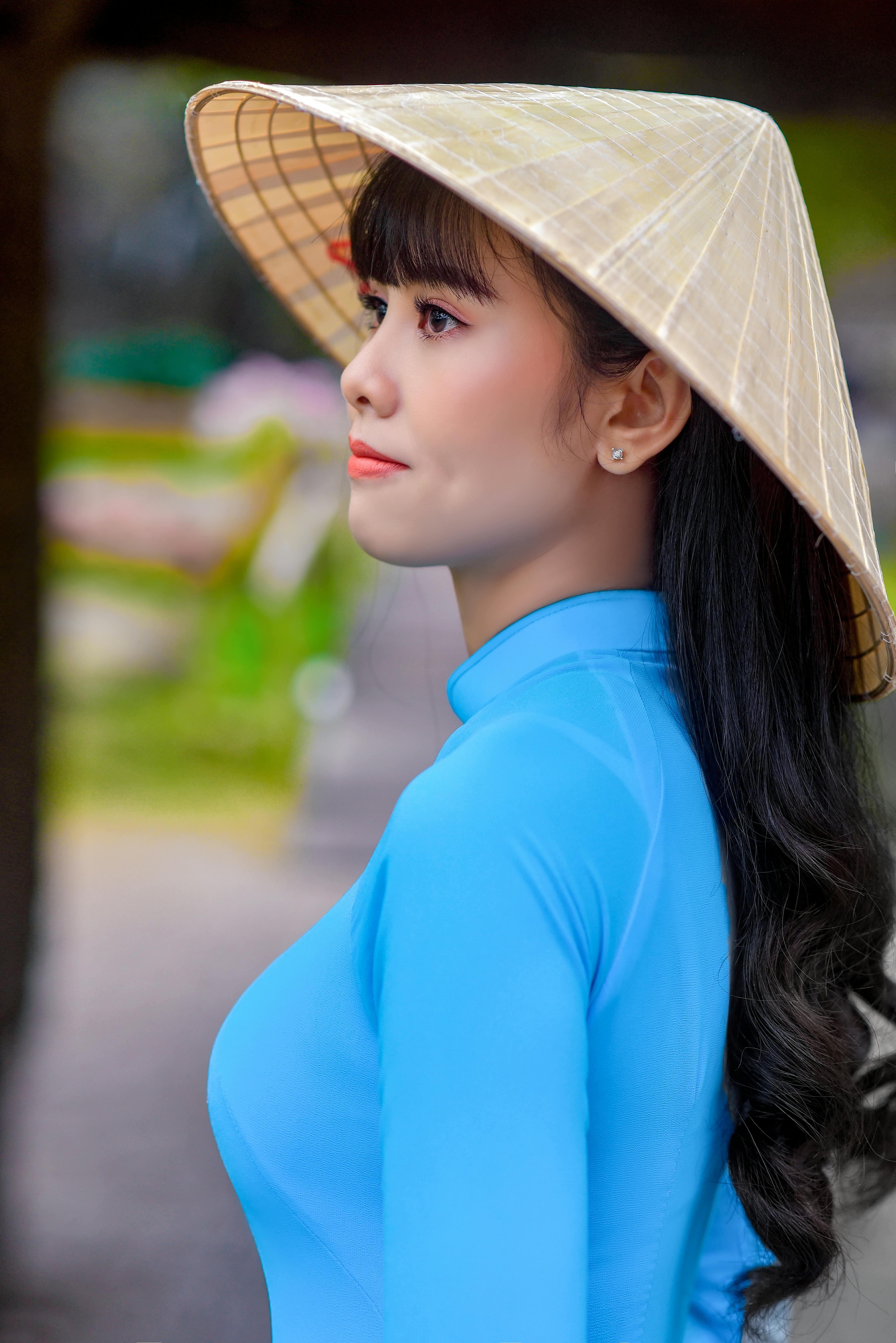 Profile portrait of an Asian woman wearing a traditional conical hat and blue dress outdoors.