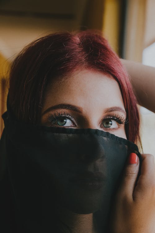 Free Beautiful young female covering pretty face with black translucent veil and looking at camera in room Stock Photo