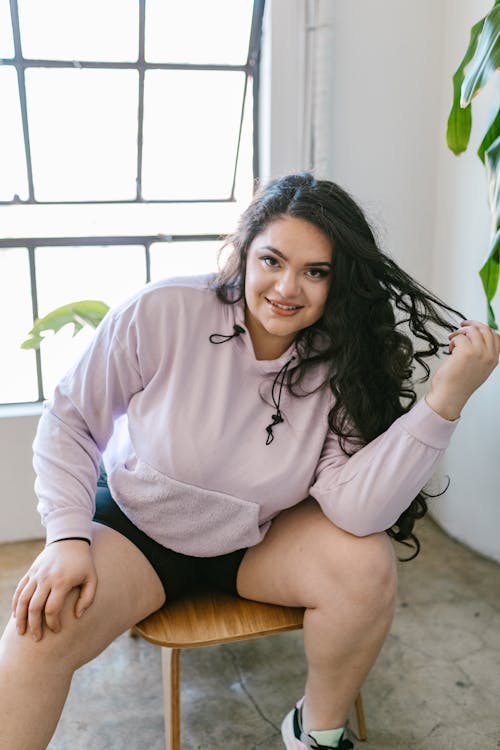 Woman in a Lavender Hoodie Sitting on a Chair while Touching Her Hair