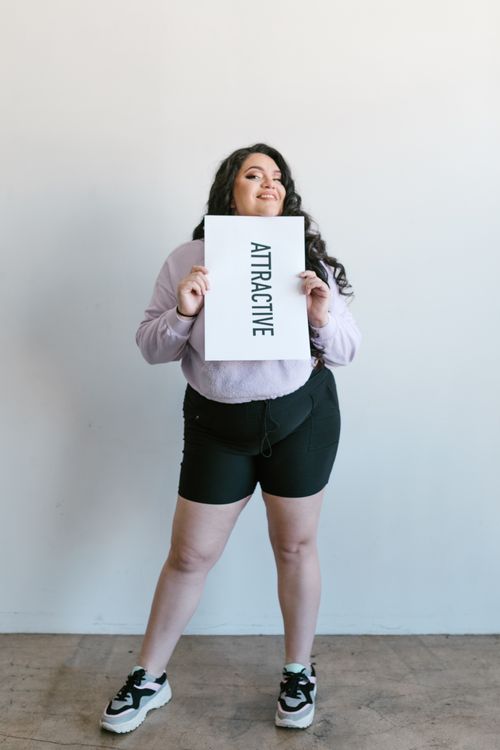 Free Plus-size woman expresses confidence holding an 'ATTRACTIVE' placard indoors. Stock Photo