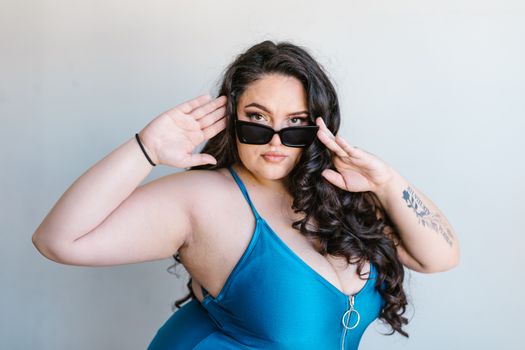 A stylish plus-size woman posing confidently in a blue swimsuit with sunglasses, exuding self-love.