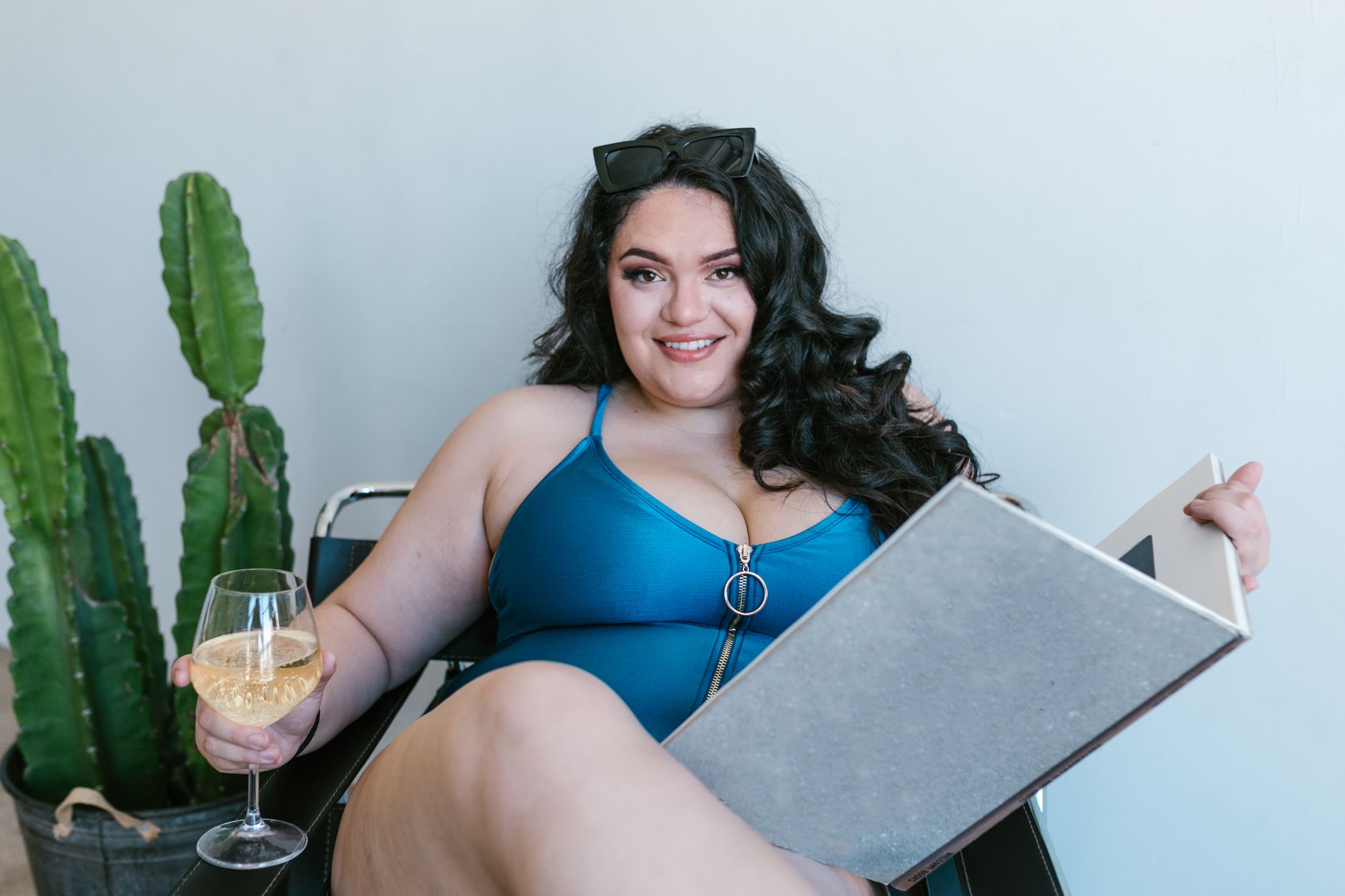 Plus-size woman in a blue bathing suit enjoying a book and wine indoors with a cactus plant.