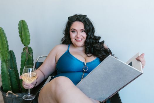 Plus-size woman in a blue bathing suit enjoying a book and wine indoors with a cactus plant.