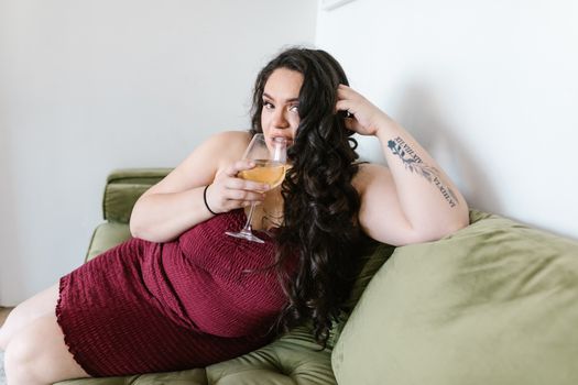Curvy woman with arm tattoo enjoying a glass of wine while lounging on a couch, embracing body positivity.