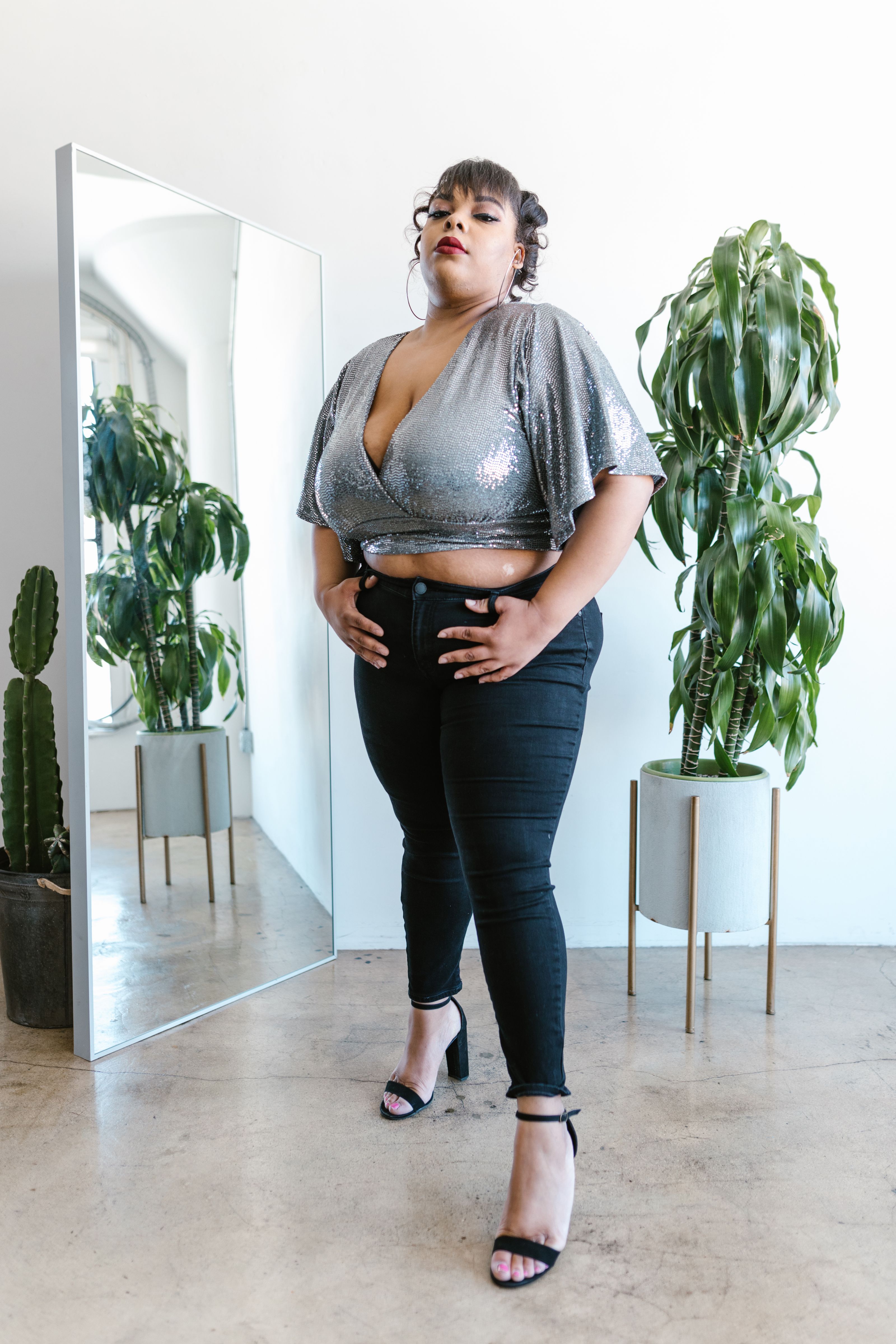 Mulher confiante plus size posando em ambiente interno com roupas da moda, plantas e espelho.