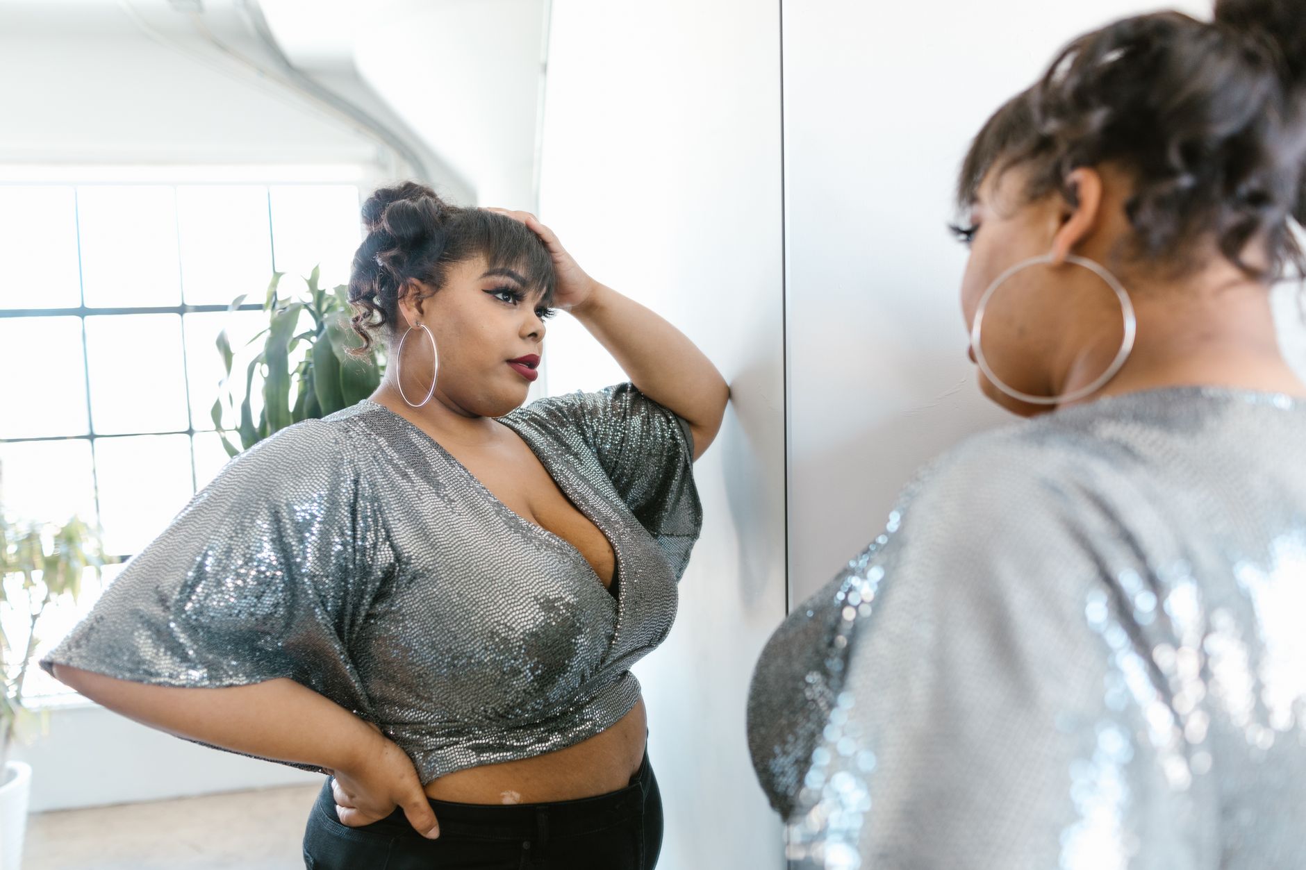 A confident plus size woman in a sparkling top poses before a mirror, embracing self love in style.